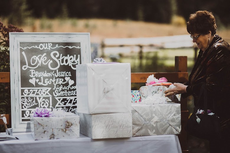 middle aged woman puts a gift down next to a pile of other wedding gifts at an outdoor wedding, silver and white packages