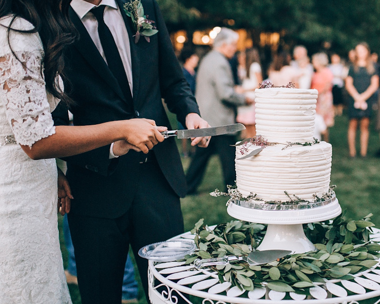 young bride and groom cut a two-tiered white wedding cake together at an outdoor wedding
