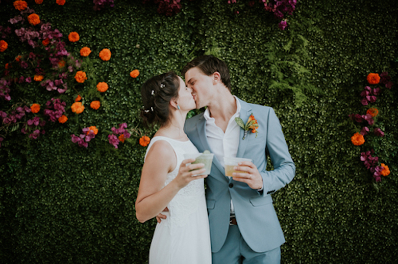 newly-wed couple kissing in front of backdrop made of greenery and flowers