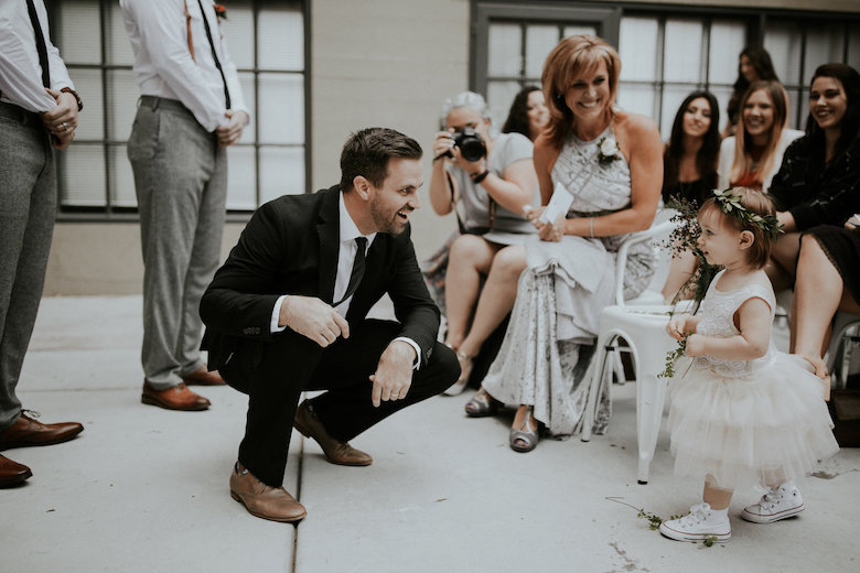 groom crouches down to look at the sweet flower girl, everyone looks on happily