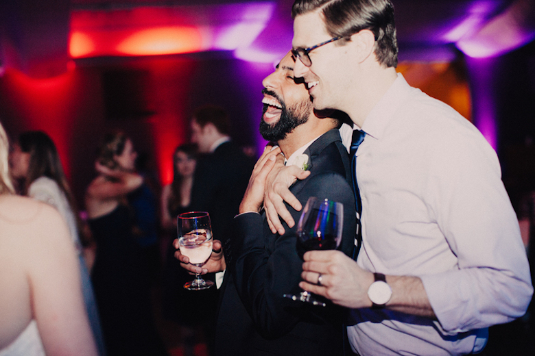 two good friends at a wedding party holding glasses of wine, embracing and laughing