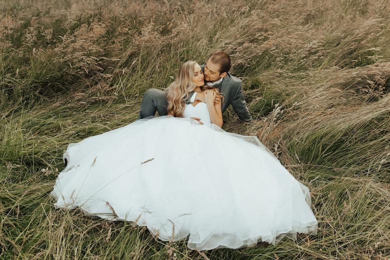 bride and groom sitting in tall grass holding each other