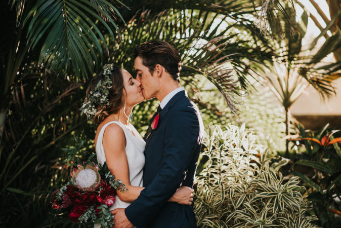 young newlyweds share a kiss under tropical foliage, outdoors sunny day