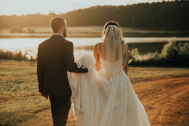 bride and groom walking in a field with a lake at sunset