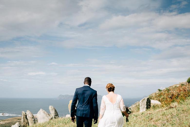 bride and groom walking near the ocean along a hill with large stones and holding hands while the bride carries a bouquet