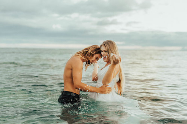 shirtless groom and bride in wedding dress stand in the ocean laughing