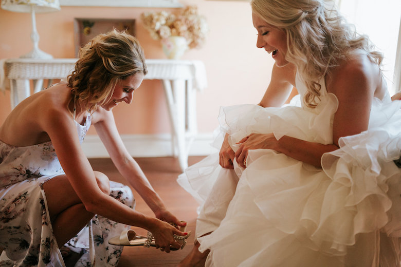 Bridesmaid putting the shoes on the bride, indoors in a sunny room, all smiles