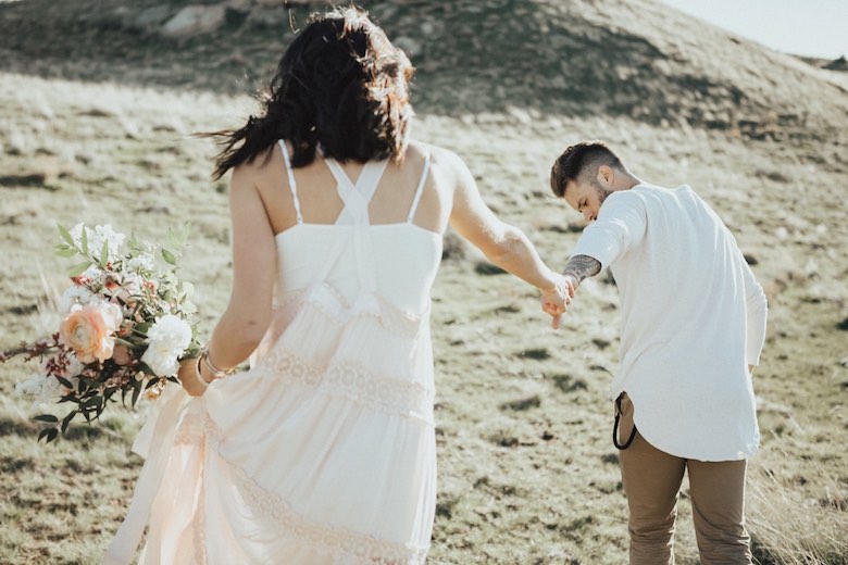 groom and bride in wedding dress holding bouquet walking along the beach holding hands